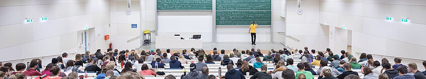 Studierende (Rückenansicht) im Hörsaal mit Vortagenden an der Tafel